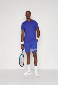 Mužský tenista v modrém tričku a šortkách, držící tenisovou raketu, v bílých zápěstních bandážích, ponožkách a teniskách na jednobarevném pozadí.