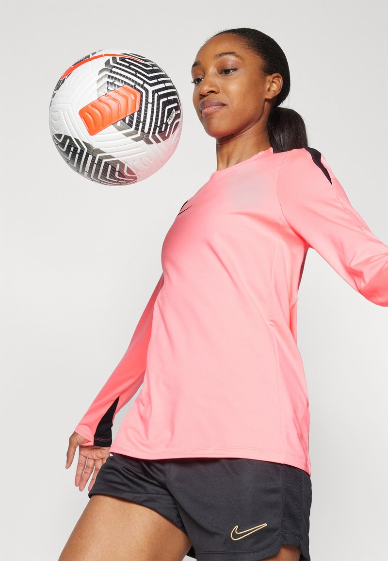 T-shirt de sport à manches longues, rose vif avec des accents noirs, assorti à un short noir. Un ballon de football à motifs flotte devant.