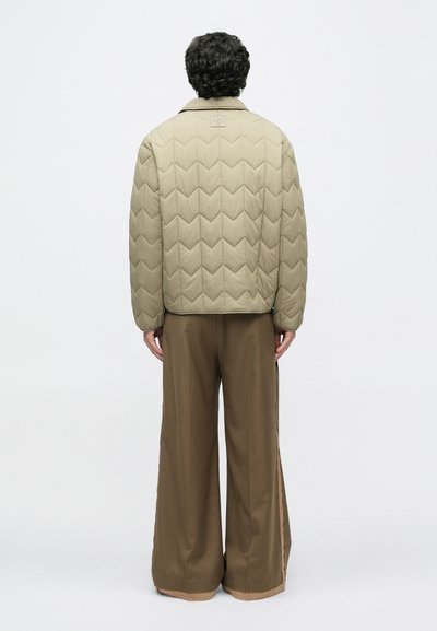 Μπεζ καπιτονέ μπουφάν με σχέδιο ζιγκ-ζαγκ, ραφές ώμων και ετικέτα στο πίσω μέρος, συνδυασμένο με φαρδιά καφέ παντελόνια με περιφραγμένα άκρα.