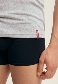 Szara bawełniana koszulka z czerwoną metką u dołu, zestawiona z dopasowanymi czarnymi szortami. Materiał wydaje się gładki i elastyczny. Ręka oparta na udzie.