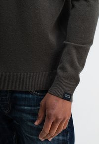 Dunkelgrüner Strickpullover mit gerippten Bündchen und Saum. Auf dem Bündchen befindet sich ein Etikett. Eine Hand mit sichtbarem Narben liegt auf der Taille nahe an der Jeans.