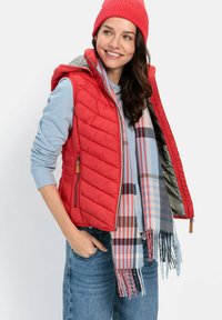 Rød quilted vest med hætte, lagdelt over en lyseblå hoodie og et flerfarvet ternet tørklæde. Denim jeans fuldender outfittet.