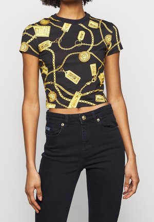 Černé oříznuté tričko se vzory zlatých řetízků a cedulek, doplněné o černé kalhoty s vysokým pasem, nošené osobou stojící před jednobarevným pozadím.