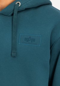 Sinine kapuutsiga jakki pehme tekstuuriga, millel on nöör ja ristkülikukujuline logopatch, millel on tume tekst "ALPHA INDUSTRIES".