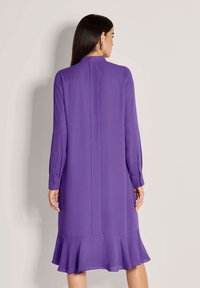 Frau mit langen dunklen Haaren, die ein lilafarbenes Langarmkleid mit geknöpften Manschetten und einem gerafften Saum trägt, mit dem Rücken zu einer einfarbigen Hintergrund.