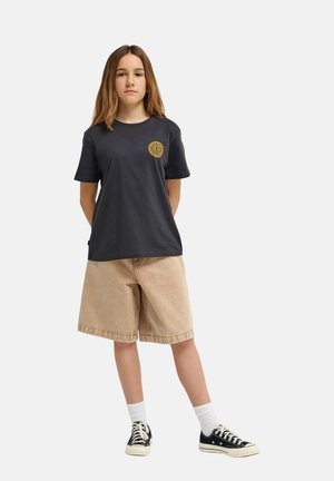 Czarny bawełniany t-shirt z żółtym okrągłym nadrukiem na lewej piersi, w połączeniu z beżowymi szortami dżinsowymi. Model nosi czarne trampki i białe skarpetki.