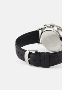 Bracelet de montre en cuir noir avec surpiqûres contrastées, quincaillerie en argent et fond en acier inoxydable poli. Présente une boucle de fermeture.