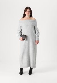 Siva pletena obleka brez naramnic z dolgimi napihnjenimi rokavi in rebrastim ovratnikom. Obuta v črne pete in drži črno torbico.