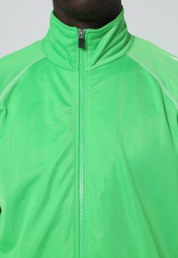 Gros plan d'une personne portant une veste zippée vert vif à col haut avec des coutures blanches le long des épaules, sur un fond clair.