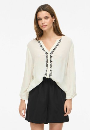 Femme portant un chemisier ample crème orné d'une bordure noire brodée, associé à un short noir taille haute, sur fond blanc.