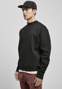 Svart sweatshirt med rund hals, ribbede mansjetter og nederkant. Båret over en lysefarvet skjorte, kombinert med burgunderbukser.
