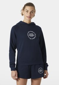 Sudadera navy con un gran logotipo gráfico circular en blanco. Material suave, bolsillo canguro y puños de canalé. Combinada con pantalones cortos navy a juego.