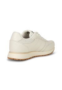 Světlé krémové nízké tenisky s tkaničkami, texturovanou boční částí a hnědou gumovou podrážkou, zobrazené z zadního úhlu.