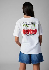 Vit grafisk t-shirt med röda körsbär och texten "Delizioso Fresh & Tasty" på ryggen, kombinerad med jeansshorts.