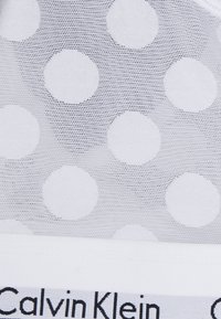 Tecido branco transparente com grandes bolinhas espaçadas uniformemente; rotulado com "Calvin Klein" em letras pretas na parte inferior.