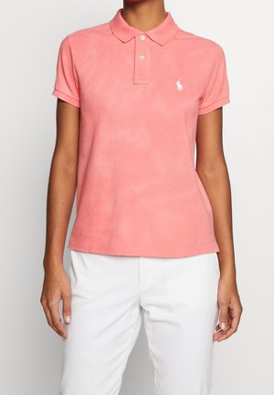 Kobieta ubrana w koralowo-różową, krótko rękawową koszulkę polo z białym haftowanym logo oraz białe spodnie, stojąca na jasnym, jednolitym tle.