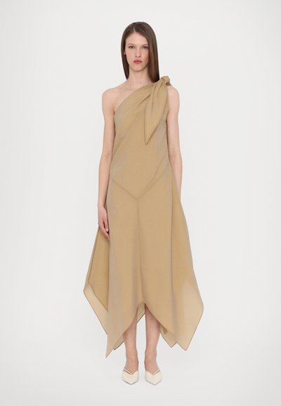 Asymmetrisches, einseitig schulterfreies Kleid in hellem Beige, aus weichem, fließendem Stoff mit einer Knotendetail am Schulterbereich. Kombiniert mit spitzen weißen Schuhen.