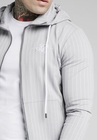 Lichtgrijze zip-up hoodie met verticale witte strepen, voorzakken en een wit logo. Inclusief een capuchon met trekkoord.