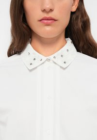 Camicia bianca con colletto strutturato adornato da strass e cuciture decorative. Presenta una chiusura con bottoni sul davanti e una forma di colletto classica.