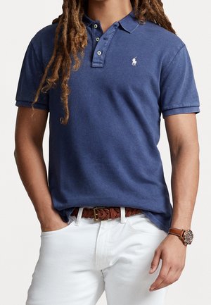 Homme portant un polo bleu avec un logo blanc, un pantalon blanc, une ceinture tressée marron et une montre-bracelet marron avec un bracelet en cuir, les mains dans les poches.