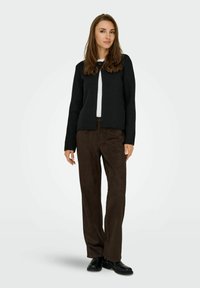 Schwarze Strickjacke mit runder Knopfleiste, über einem weißen Oberteil und dunkelbraunen Wildlederhosen getragen, kombiniert mit schwarzen Schuhen.
