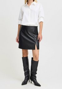 Vrouw in een witte blouse met knopen, gestoken in een zwarte leren mini-rok met een split aan de voorkant, gecombineerd met zwarte kniehoge haklaarzen.