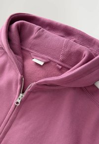 Ružová mikina na zips s ribbed vnútornou kapucňou, jemným flísovým podšívkou a kovovým zipsom. Obsahuje štítok na výstrihu a hladký povrch.