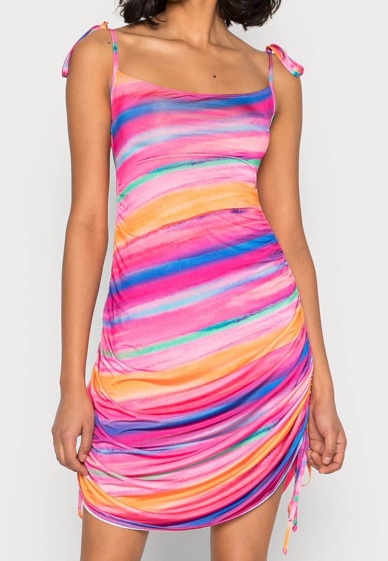 Femme portant une mini-robe sans manches froncée avec des bretelles à nouer, rayures horizontales en aquarelle rose vif, orange, bleu et vert.