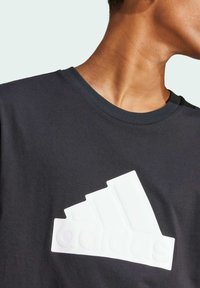 La maglietta nera presenta un grande logo Adidas bianco in gomma sul petto, con un classico scollo rotondo e una morbida texture del tessuto.