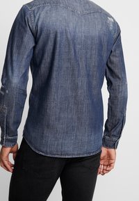 Camicia in denim blu scuro con maniche lunghe, leggera usura sulle spalle, colletto con bottoni, design aderente e orlo curvo sul retro.