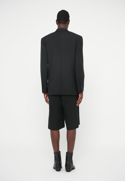 Μαύρο προσαρμοσμένο σακάκι με δομημένους ώμους, συνδυασμένο με μαύρες βερμούδες. Φορεμένο με μαύρες μποτίνες μέχρι τον αστράγαλο. Απαλή υφή, ελάχιστες λεπτομέρειες.