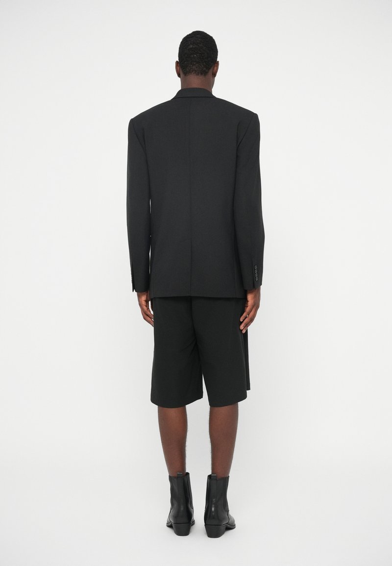 Sort skræddersyet blazer med strukturerede skuldre, parret med sorte shorts. Båret med sorte ankelstøvler. Glat tekstur, minimal detaljer.