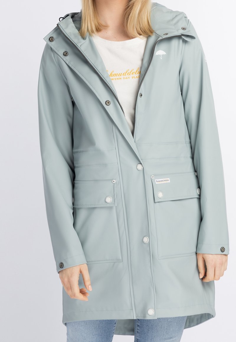 Femme portant un imperméable à capuche gris clair avec des boutons pression, plusieurs poches avant, et un t-shirt blanc en dessous.