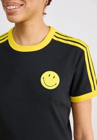 Black t-shirt with a yellow round neckline and yellow stripes on the sleeves. Features a yellow smiley face embroidered on the chest.
