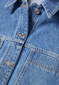 Section d'une veste en denim bleu clair montrant le col, les boutons-pression métalliques, la poche avant et les détails de couture orange.