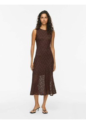Femme aux longs cheveux bouclés portant une robe midi marron en dentelle sans manches et des talons ouverts noirs sur fond blanc uni.