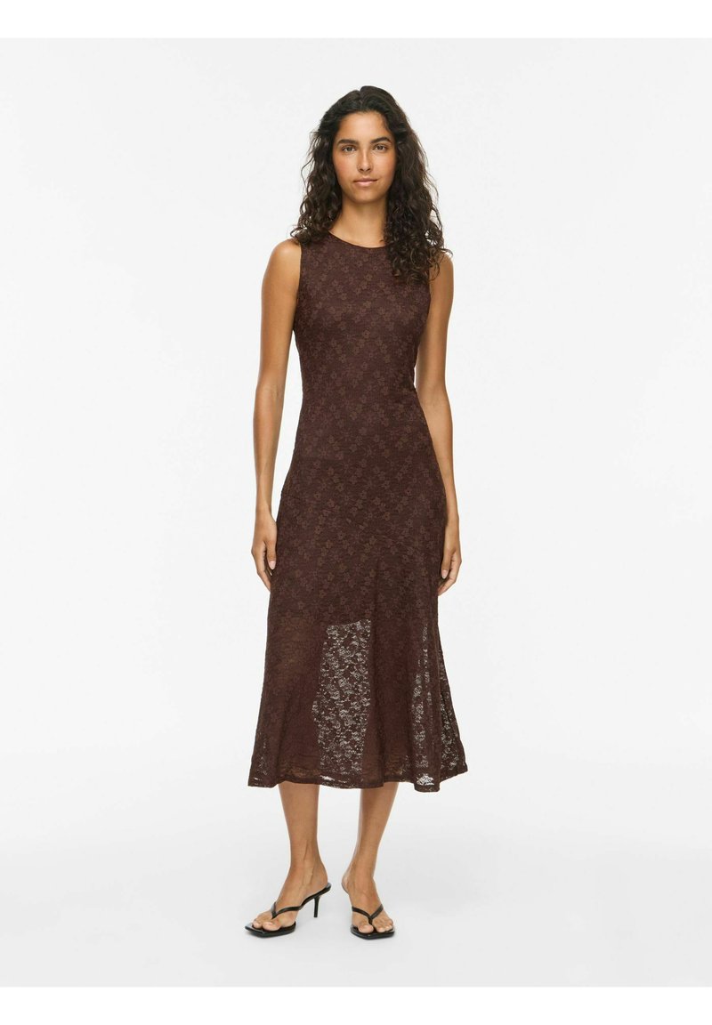 Femme aux longs cheveux bouclés portant une robe midi marron en dentelle sans manches et des talons ouverts noirs sur fond blanc uni.