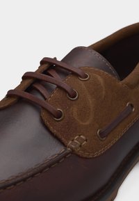 Καφέ δερμάτινο και σουέτ παπούτσι τύπου boat με στρογγυλά μεταλλικά ρουμπιλάκια και καφέ δερμάτινα κορδόνια, κοντινή λήψη στη ραφή μπροστά και στο πλάι.