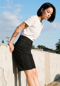 Fiatal nő fehér pólóban és fekete térdig érő rövidnadrágban, háttal egy fehér betonfalnak dőlve, szabadban, kék ég alatt.