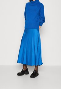 Sinine kootud kõrge kaelusega sviiter tekstuursete varrukatega, koos sinise satiinist midi-seelikuga. Mustad massiivsed mokassiinid täiendavad riietust.