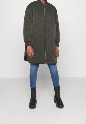 Personne portant une longue veste matelassée vert foncé avec une fermeture éclair à l’avant, un jean skinny bleu clair et des bottines noires, debout devant un fond uni.
