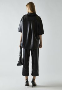 Camisa oversized de cuero negro con mangas cortas, combinada con pantalones acampanados a juego. Los accesorios incluyen un bolso negro y zapatos de tacón alto.