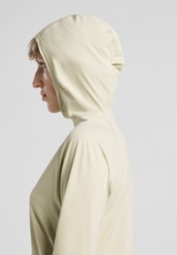 Kapuzensweatshirt aus weichem, blassem Gelb. Verfügt über eine glatte Textur und eine enganliegende Passform, mit sichtbaren Nähten entlang der Schultern und Seiten.