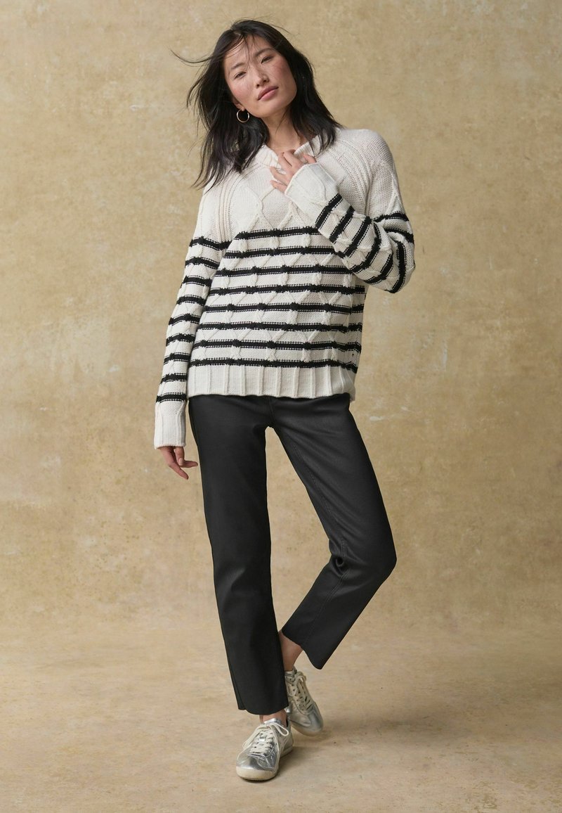 Creme trui met zwarte horizontale strepen, textuurpatroon; gecombineerd met zwarte slim-fit broek en zilveren sneakers. Effen achtergrond.