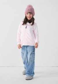 Felpa con cappuccio rosa chiaro con cerniera, jeans cargo blu larghi con tasche, pantofole grigie e berretto rosa lavorato a maglia con pompon in cima.