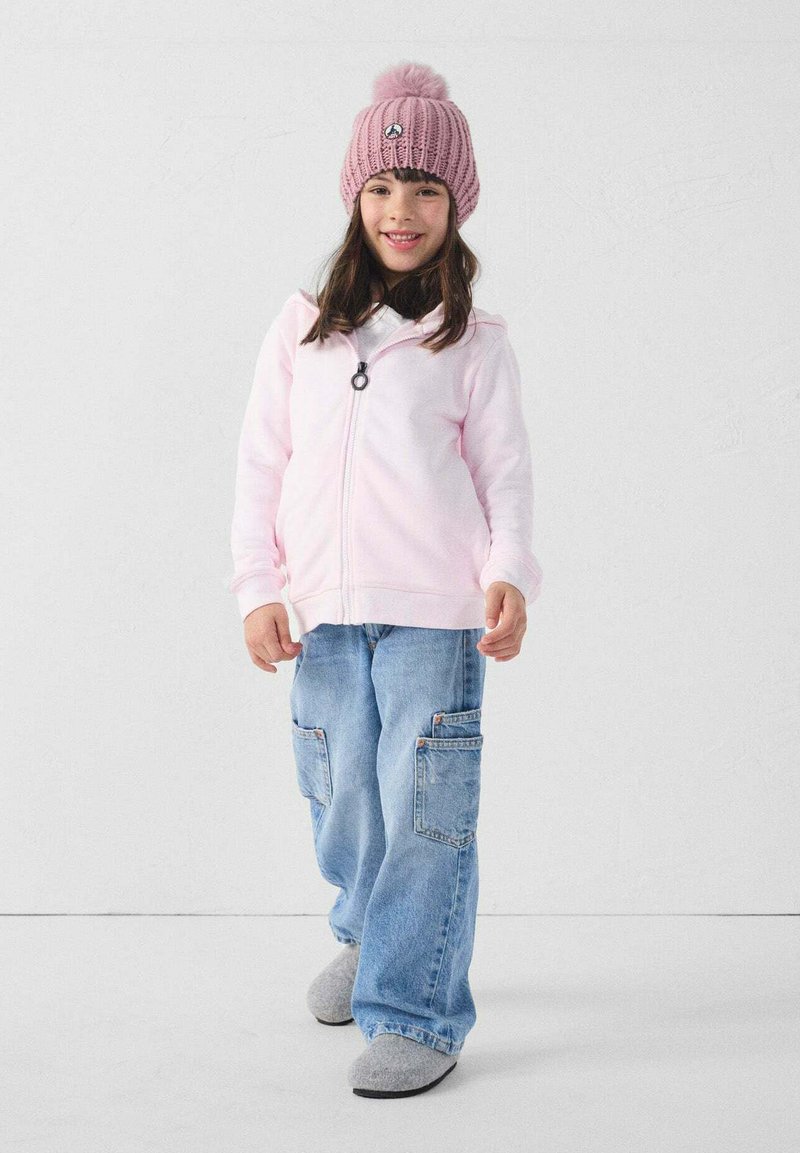 Felpa con cappuccio rosa chiaro con cerniera, jeans cargo blu larghi con tasche, pantofole grigie e berretto rosa lavorato a maglia con pompon in cima.