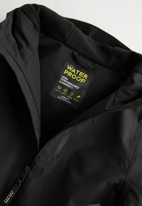 Veste noire imperméable avec doublure en polaire, étiquette indiquant des caractéristiques de haute performance et fermeture éclair partiellement ouverte.