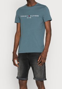 Krátký rukáv modré bavlněné tričko s kulatým výstřihem a bílým logem "TOMMY HILFIGER", spárované s ohrnutými černými džínovými šortkami.