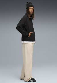 Giacca nera leggera con colletto alto e polsini elasticizzati, abbinata a pantaloni beige larghi e sneakers nere. Berretto nero con un motivo.