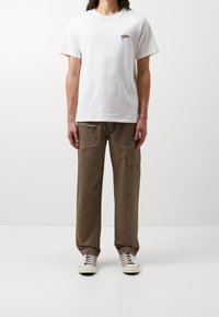 Hvid bomulds t-shirt med lille farverigt emblem på brystet, parret med løse brune cargo-bukser med lommer og snor. Lyse sneakers.